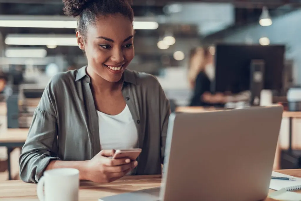 A seated woman smiles, with smartphone in hand, while she looks at laptop screen.