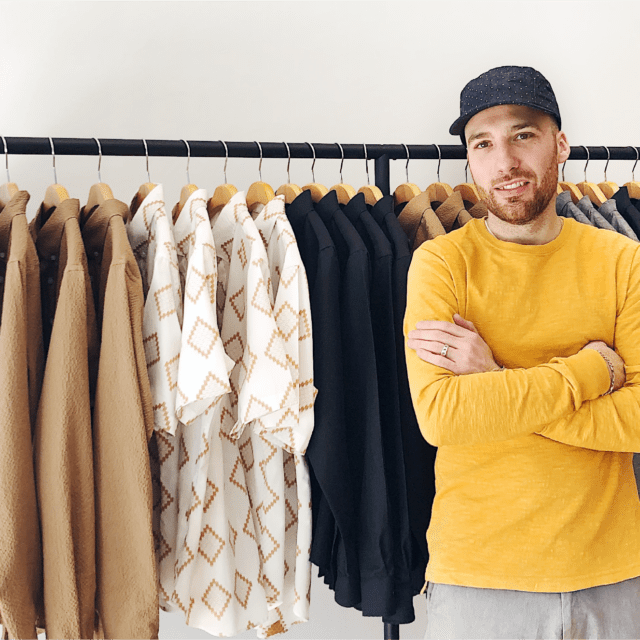 A smiling Matteo wearing a yellow sweater and ballcap stands in front of a clothing rack.