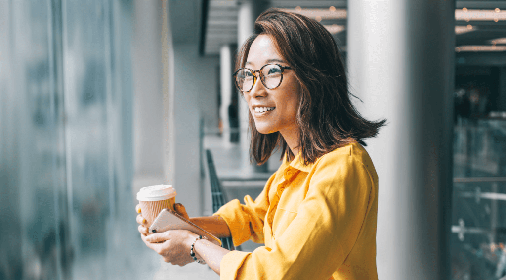 Une femme souriante, café et cellulaire à la main, regarde par la fenêtre.