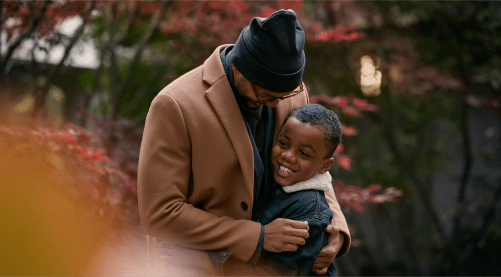 Picture of a dad hugging his smiling son, surrounded by warm, autumn colours and trees.