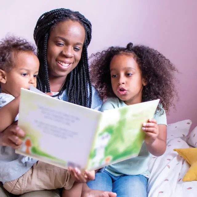 Marielle Altenor sits on a white polka dot bed reading a story book with her little son and daughter on her lap.