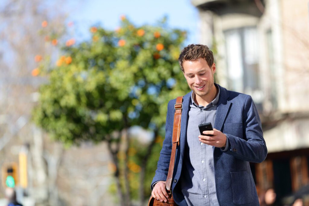 A man in a suit walking outdoors smiles as he looks at his smartphone.