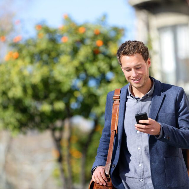 A man in a suit walking outdoors smiles as he looks at his smartphone.