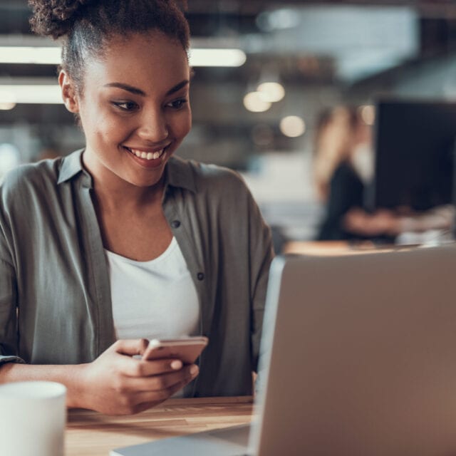 A seated woman smiles, with smartphone in hand, while she looks at laptop screen.