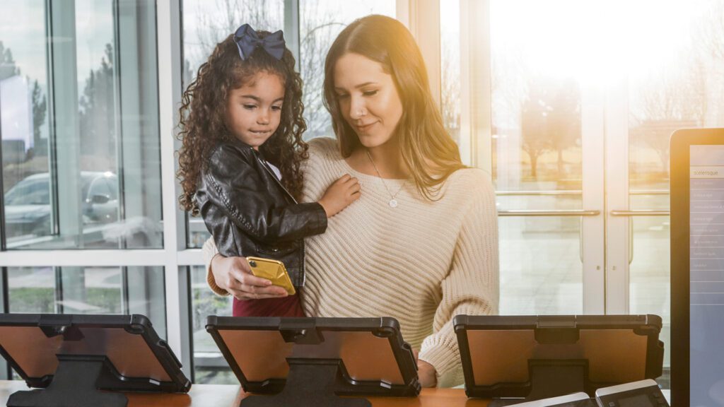 A woman holding a young girl (and a smartphone) uses a tablet at a health clinic check-in station