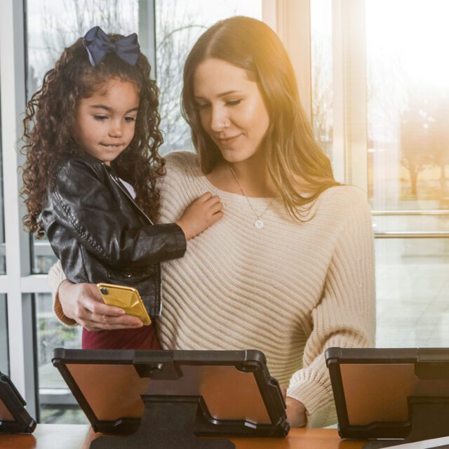 A woman holding a young girl (and a smartphone) uses a tablet at a health clinic check-in station