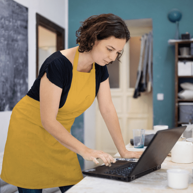 Une femme qui porte un tablier jaune utilise un ordinateur portable dans l’atelier d’une petite entreprise.