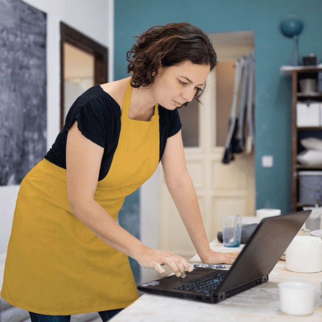 A woman wearing an ochre-coloured apron uses a laptop in a small business workshop.