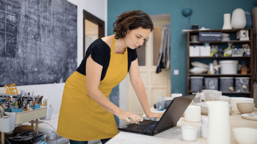 A woman wearing an ochre-coloured apron uses a laptop in a small business workshop.