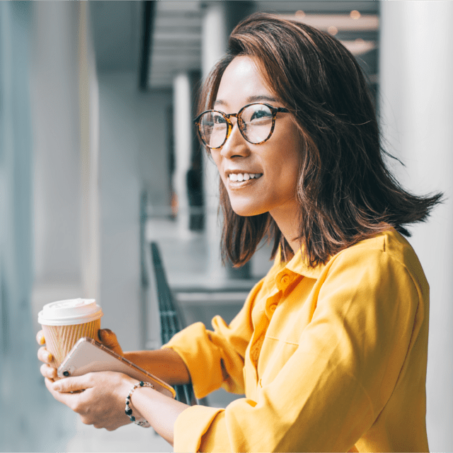 A smiling woman holding a coffee and cellphone looks out the window.