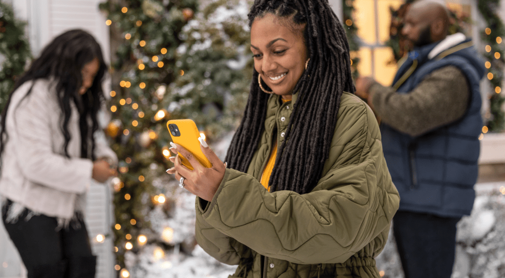 Woman in green coat looks down at her yellow phone and smiles as people decorate behind her.