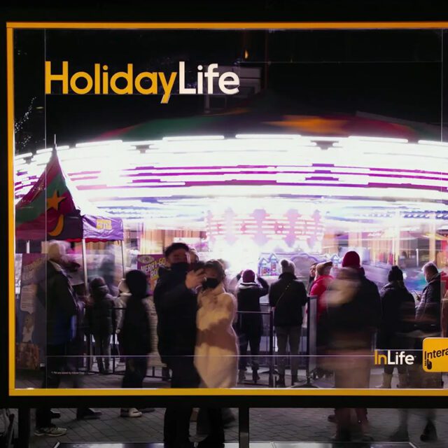 A couple taking a picture through an Interac HolidayLife transparent billboard at a busy night carnival