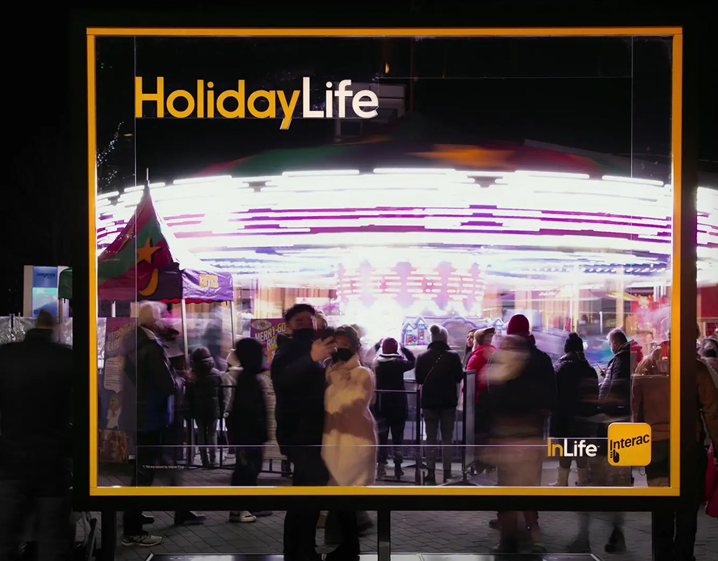A couple taking a picture through an Interac HolidayLife transparent billboard at a busy night carnival