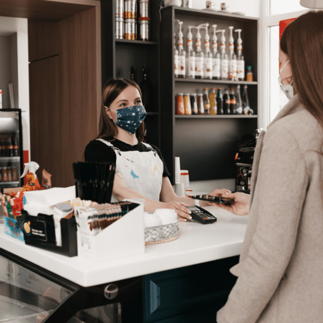 Masked woman behind café counter offers a payment terminal for a debit payment.