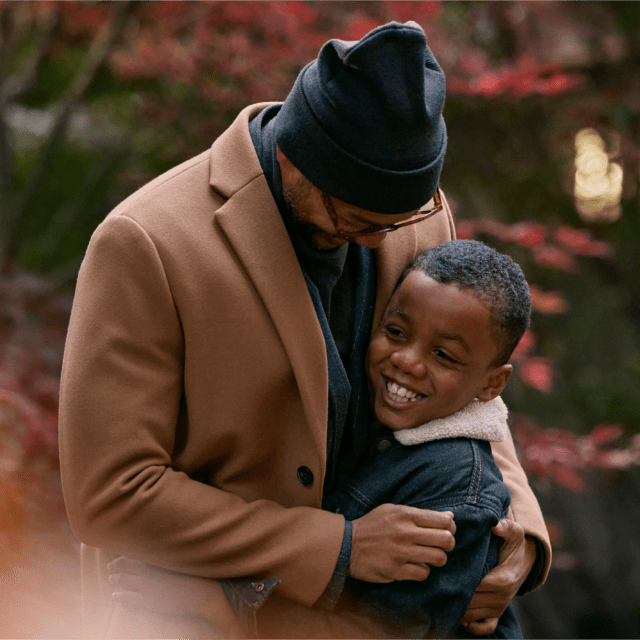 Picture of a dad hugging his smiling son, surrounded by warm, autumn colours and trees.