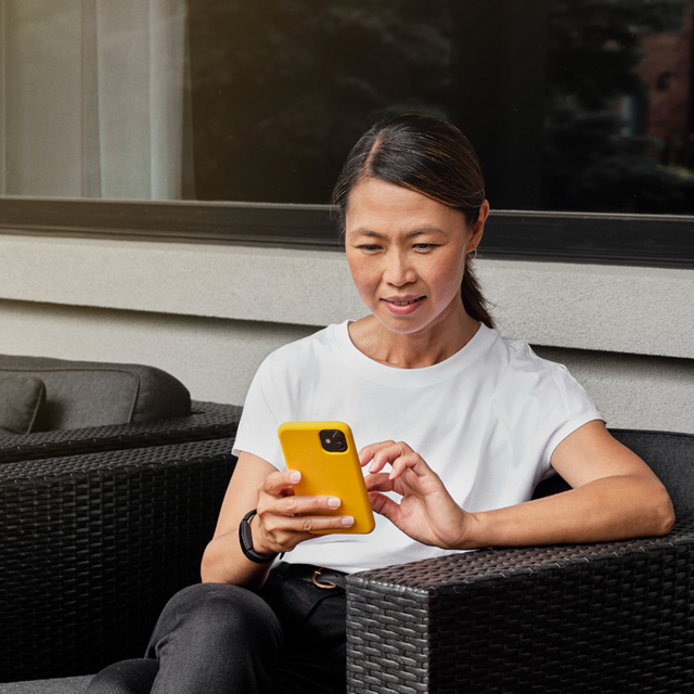 Woman sits on her deck using her cellphone to make an Interac e-Transfer for Business transaction.