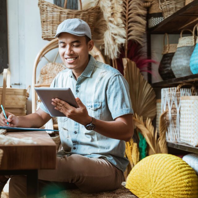 Open banking: Smiling man in a stock room full of woven baskets holds a pen, clipboard and tablet.