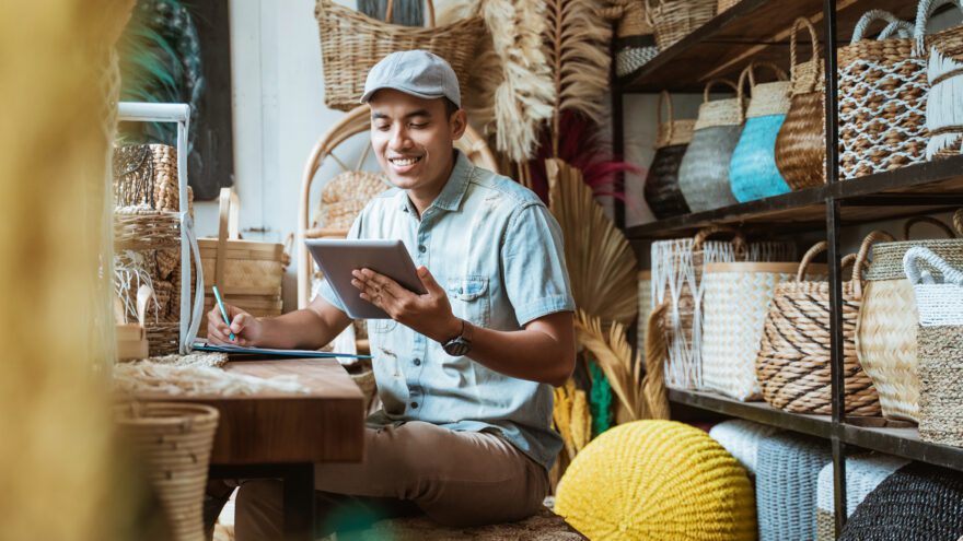 Open banking: Smiling man in a stock room full of woven baskets holds a pen, clipboard and tablet.