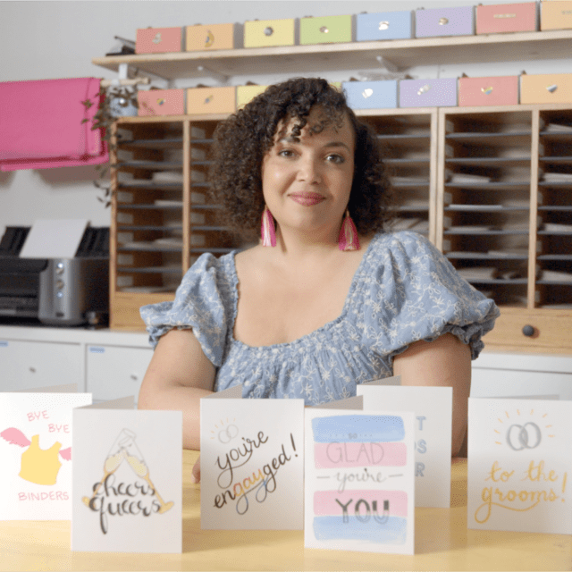 Heather sits smiling behind a desk on which her bright, pastel coloured cards are displayed.