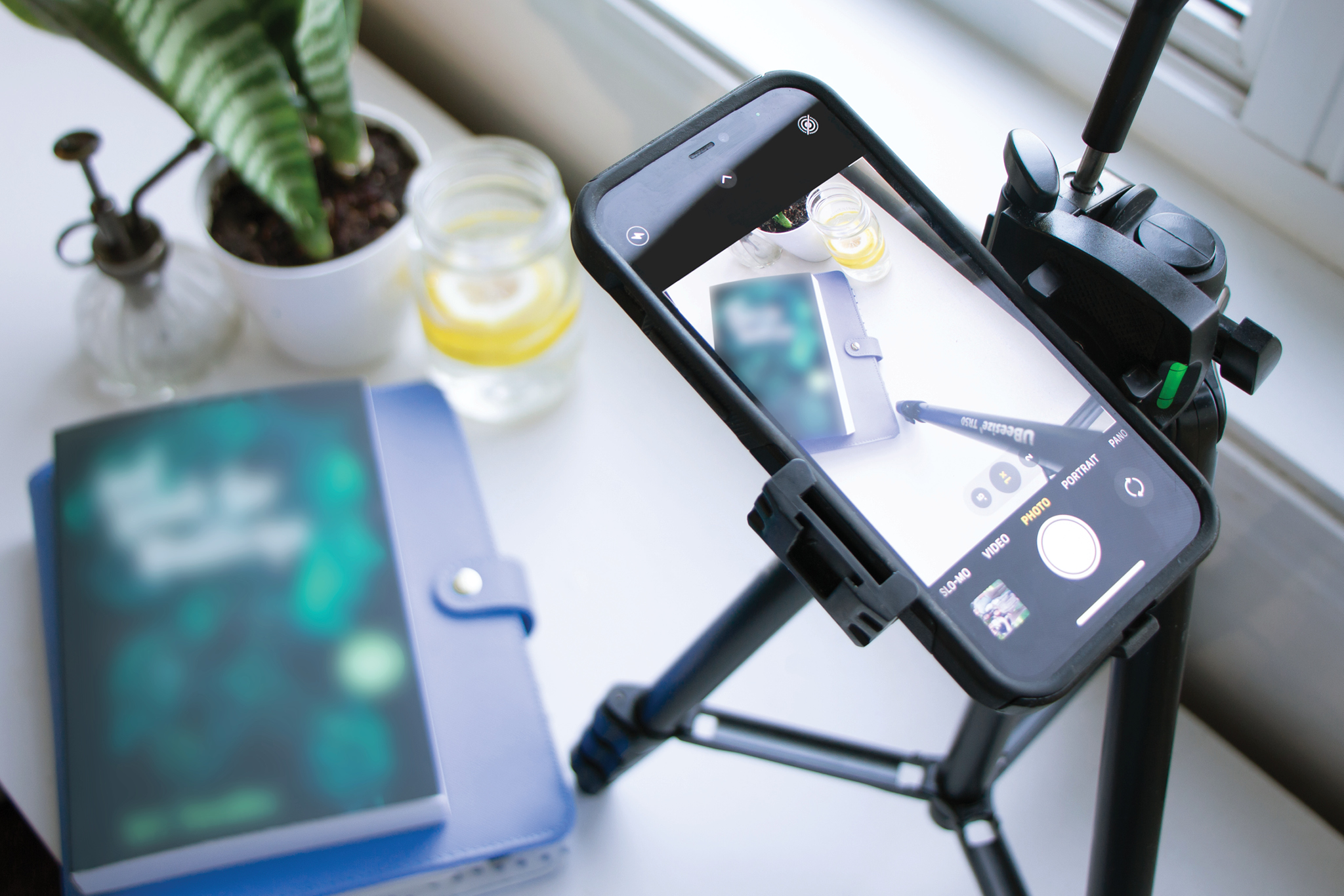 A tripod with a phone attached is shown taking a photo of a dark green book flatlay from above.