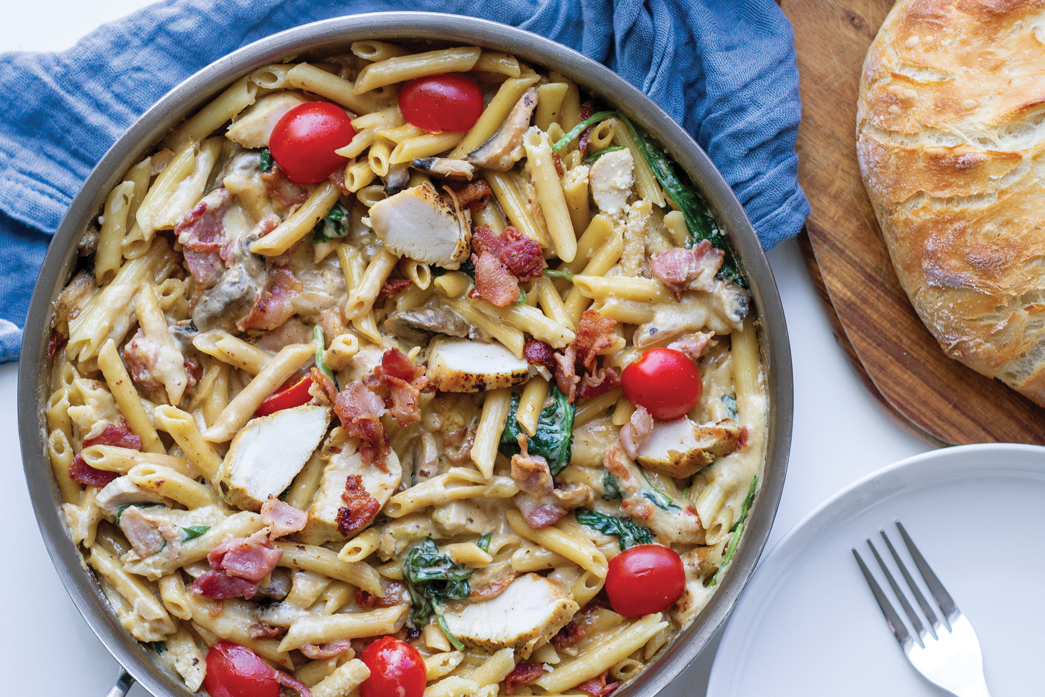 A flatlay of a colourful plate of chicken pasta with red cherry tomatoes and a round loaf of bread on the side.