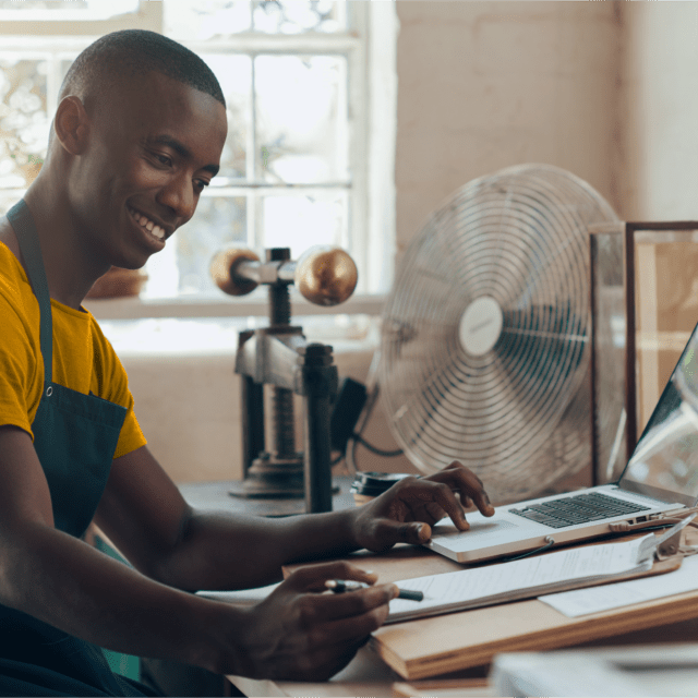 A small business owner smiling while he works on his computer and with a clipboard