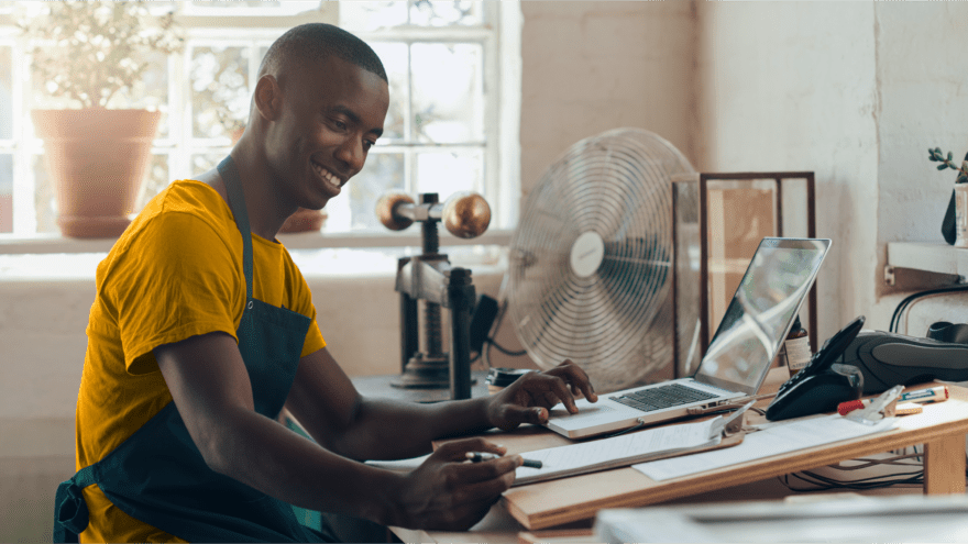 A small business owner smiling while he works on his computer and with a clipboard